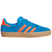 A blue suede Adidas Gazelle sneaker with bright orange stripes, a gum sole, and gold 'GAZELLE' text on the side.