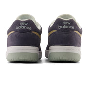 A pair of New Balance low-top sneakers shown from the back, featuring dark grey suede uppers, a white midsole, a light grey green outsole, and gold accents.