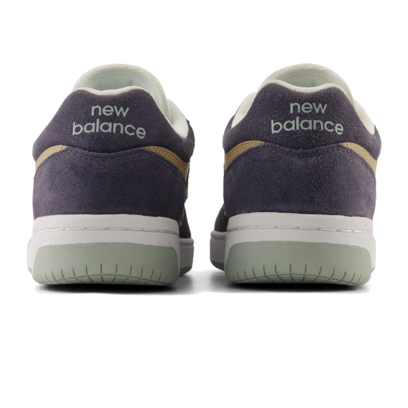 A pair of New Balance low-top sneakers shown from the back, featuring dark grey suede uppers, a white midsole, a light grey green outsole, and gold accents.